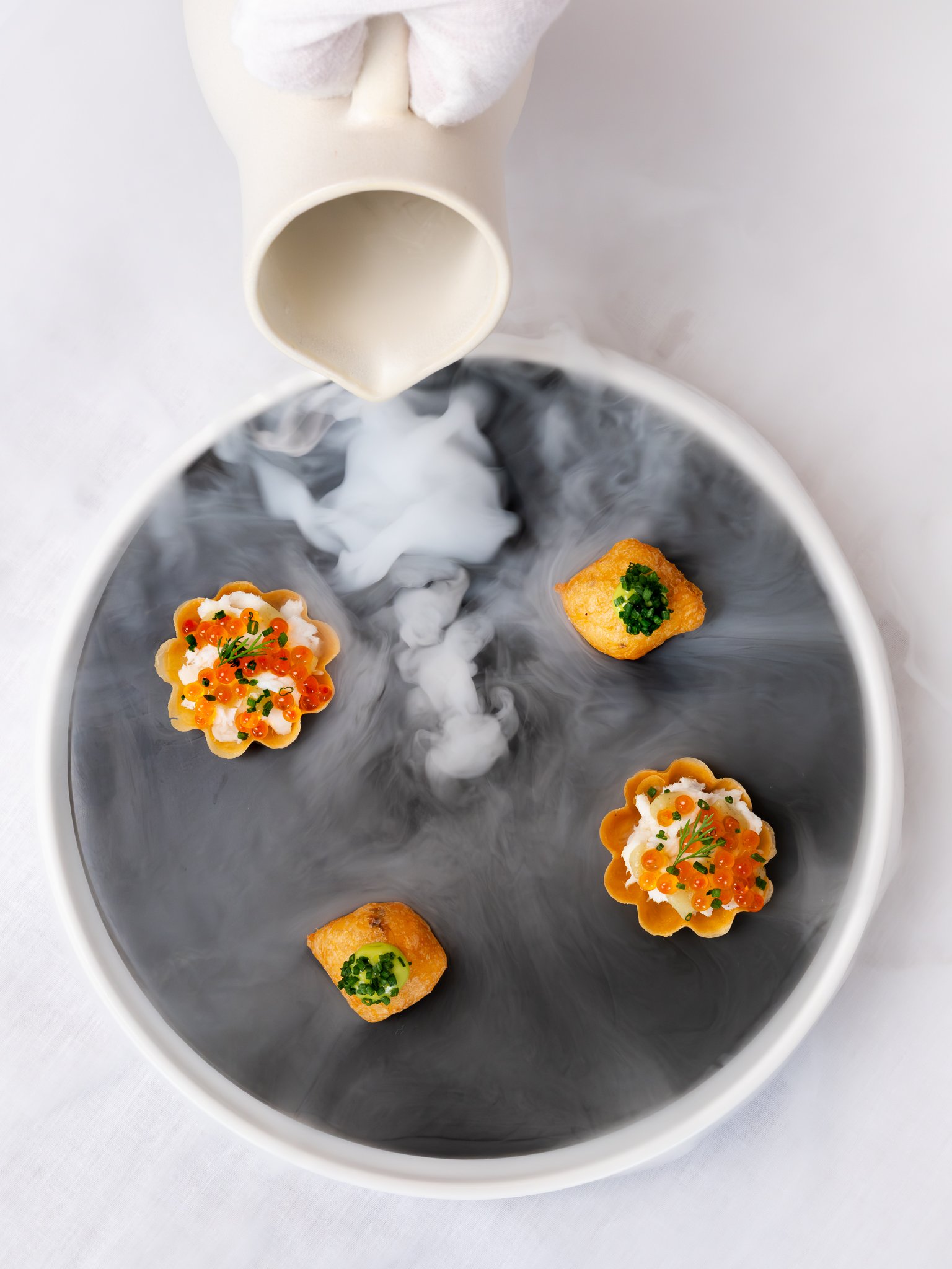 A white oval plate with two types of hors d'oeuvres: one topped with what appears to be salmon roe, a dollop of cream, and herbs, and the other a small square fried item topped with chopped chives, with a small white pitcher pouring a white sauce ont
