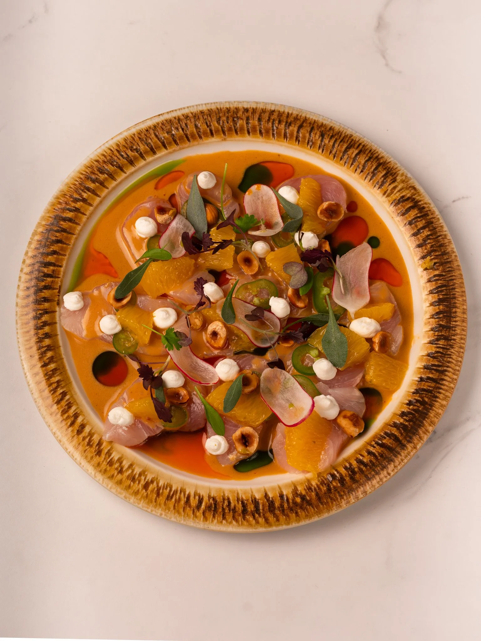 A dish of ceviche topped with microgreens, sliced radishes, orange segments, and drizzled with various sauces, served in a brown-rimmed bowl.