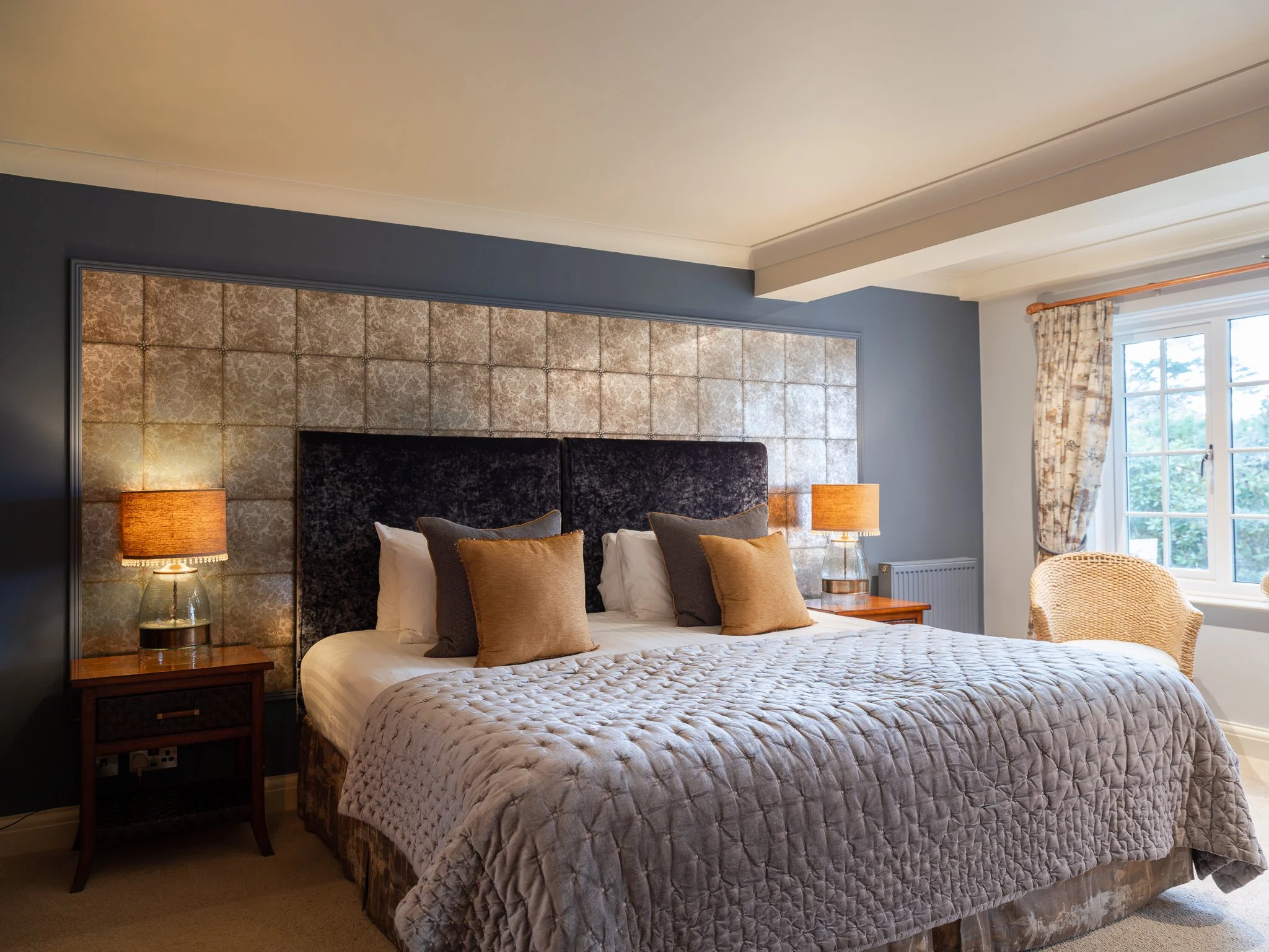 A bedroom with a large bed, dark headboard, beige quilt, four pillows, two nightstands with lamps, window with curtains, and a wicker chair.