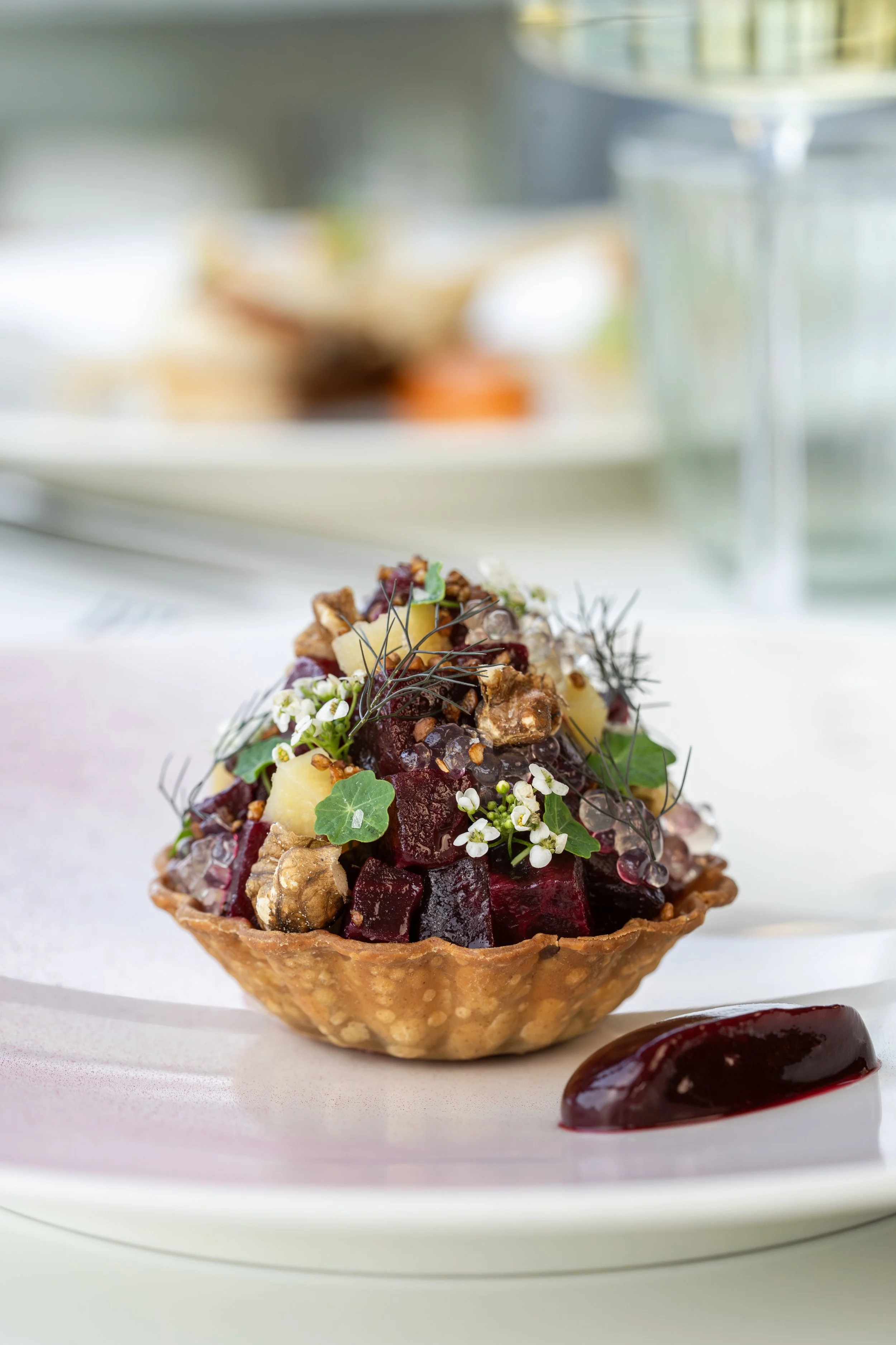 A gourmet beet salad served in a crispy tart shell, garnished with herbs, edible flowers, and nuts, with a smear of beet sauce on the side.
