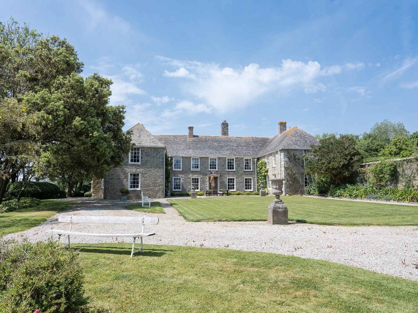 Large stone historic mansion with well-maintained lawn, pathway, trees, and sculptures in the garden under a bright blue sky with scattered clouds.