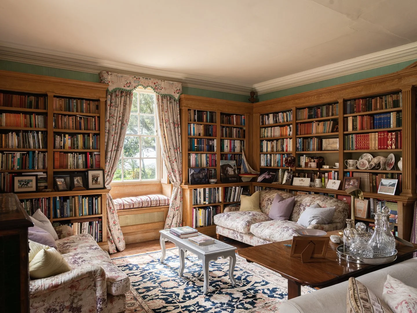 A cozy living room with wooden bookshelves filled with books, a window seat with floral curtains, a floral couch with cushions, a wooden table with glass decanters, and framed photographs, a patterned rug, and decorative plates.
