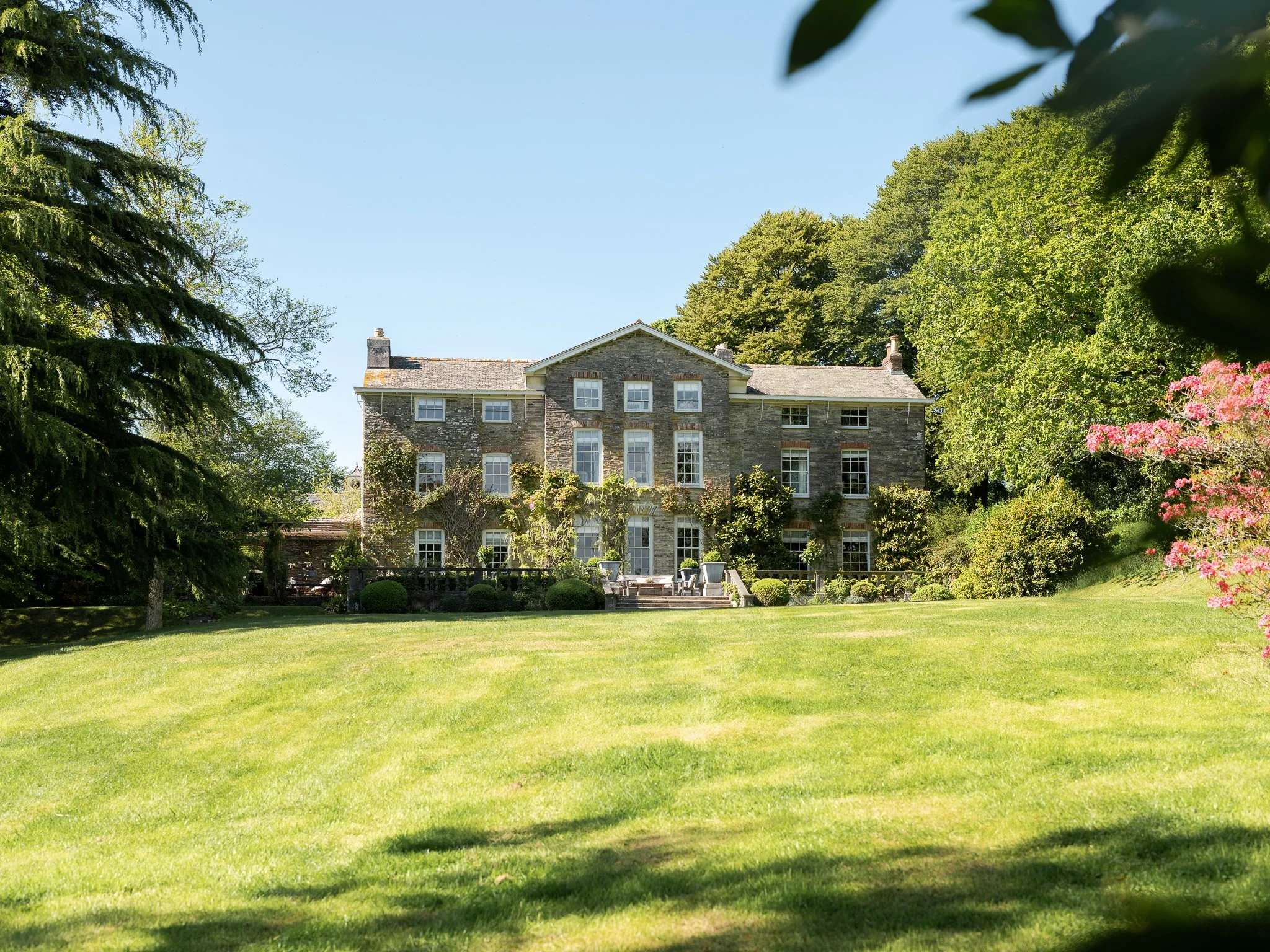 A large, historic stone house surrounded by lush green trees and a well-maintained lawn under a clear blue sky.