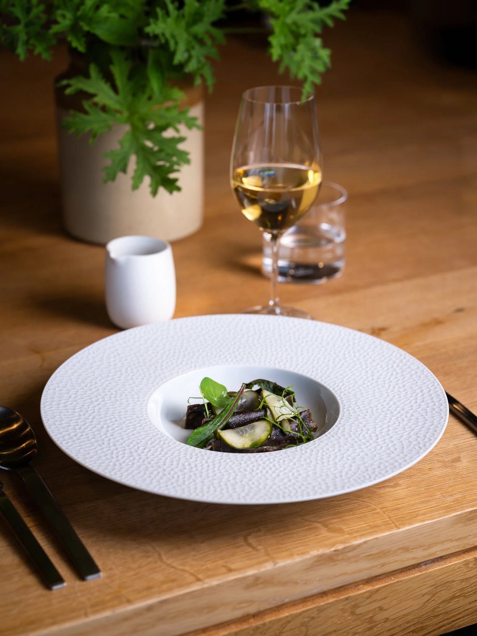 A white textured plate with a gourmet dish garnished with leafy greens on a wooden table. Behind the plate are a glass of white wine, a small white jug, a glass of water, and a potted green plant.