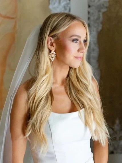 Bride with long blonde hair wearing a white dress and pearl earrings, standing indoors near a textured wall. HS Glam Makeup Bridal Portfolio