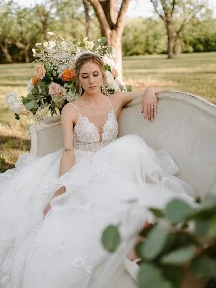A bride in a white wedding dress sitting on a vintage sofa outdoors, with a large floral arrangement behind her and a tree in the background. HS Glam Makeup Bridal Portfolio