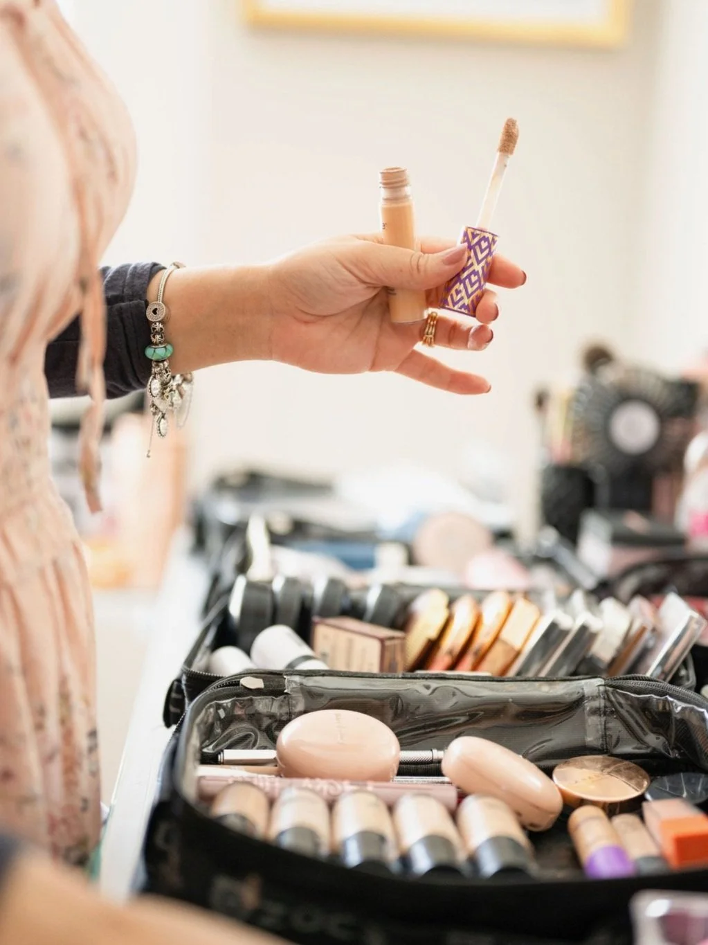 Person holding a tube of liquid foundation with an applicator brush, in front of a makeup train case filled with various makeup products. HS Glam Makeup Studio