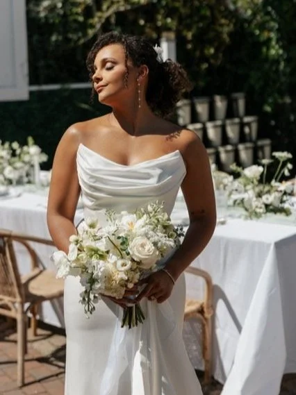 A woman in a white wedding dress holding a bouquet of white flowers, standing outside in sunlight at a wedding reception. HS Glam Makeup Bridal Portfolio