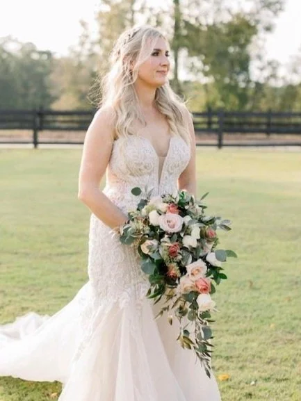 A bride with long blonde hair in a lace white wedding gown holding a large bouquet of pink and white roses and greenery, standing outdoors on a grassy field with trees and a fence in the background. HS Glam Makeup Bridal Portfolio