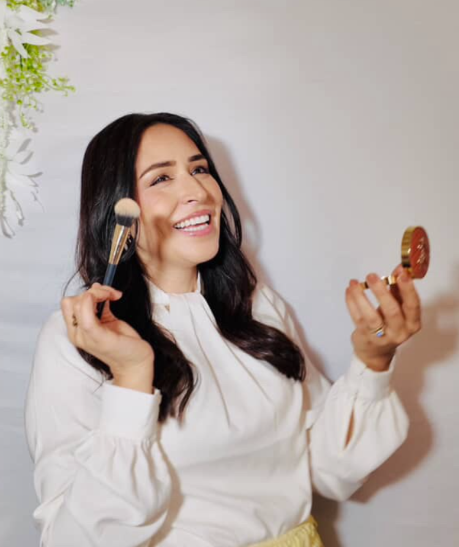 A woman with long dark hair smiling while holding a makeup brush in one hand and a small mirror in the other, dressed in a white blouse, standing against a plain wall. HS Glam Makeup Gallery