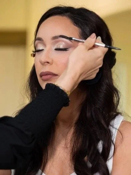 A woman with dark hair getting her makeup done, with her eyes closed, as a makeup artist applies eyeshadow with a brush. HS Glam Makeup Bridal Services
