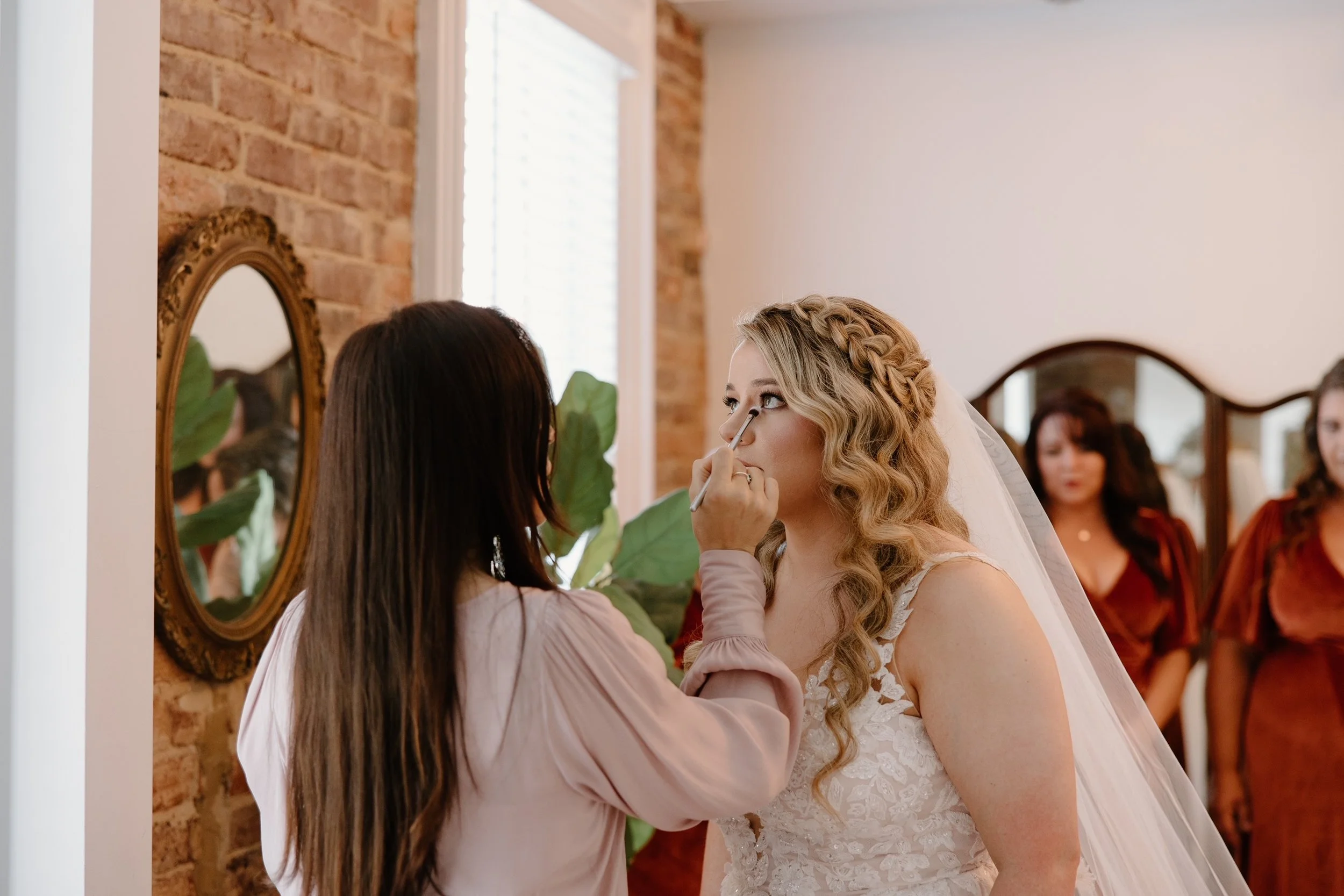 A bride in a white lace wedding dress and veil getting her makeup finished by a makeup artist, with bridesmaids in burgundy dresses in the background. HS Glam Makeup Bridal