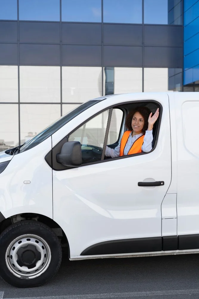 A woman with curly hair, wearing an orange safety vest, sitting in the driver's seat of a white delivery van, smiling and waving outside a modern building with large glass windows.
