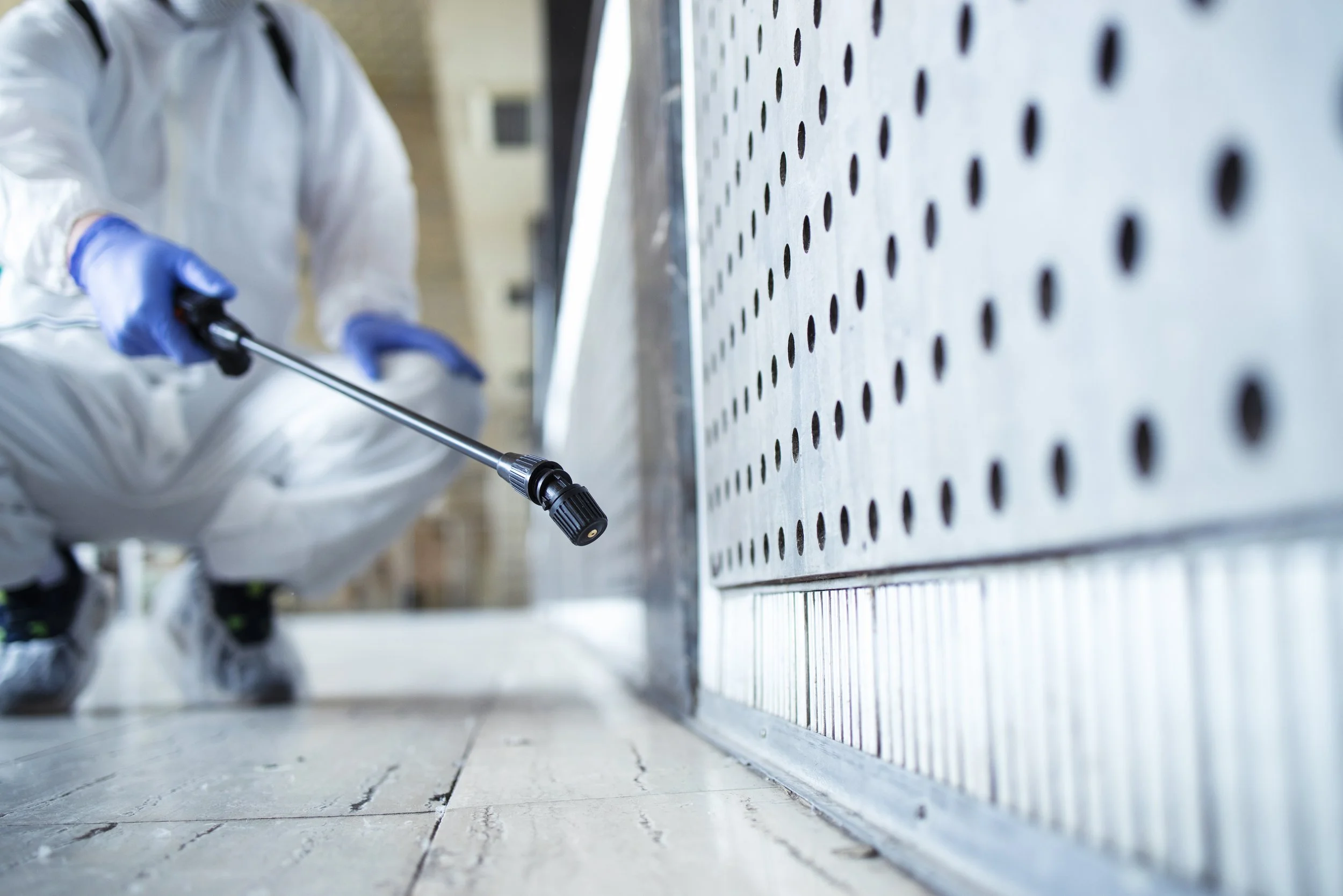 A person in white coveralls and blue gloves is kneeling and using a sprayer on the floor near a metal perforated panel.