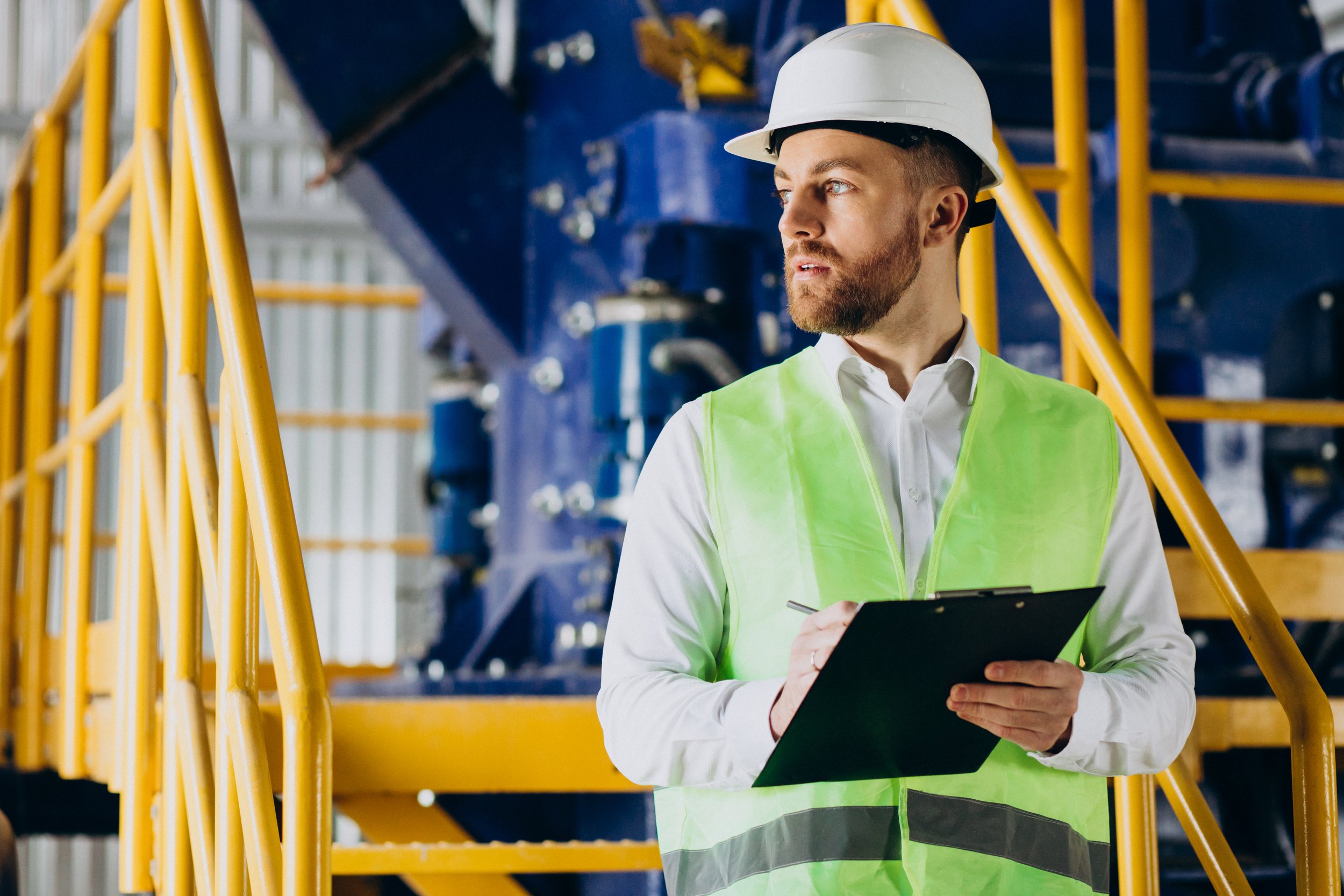 A man in a white hard hat and green safety vest holding a clipboard, standing on yellow metal stairs inside an industrial facility.