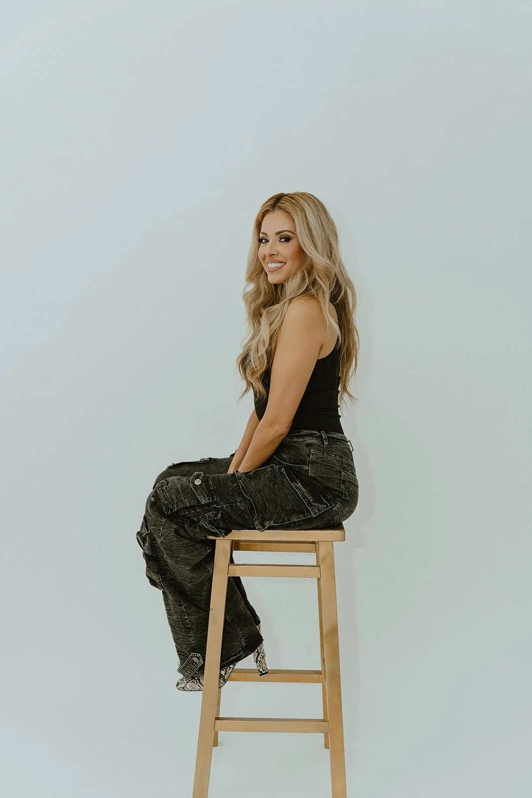 A woman with long blonde hair sitting on a wooden stool against a plain light background, smiling at the camera.