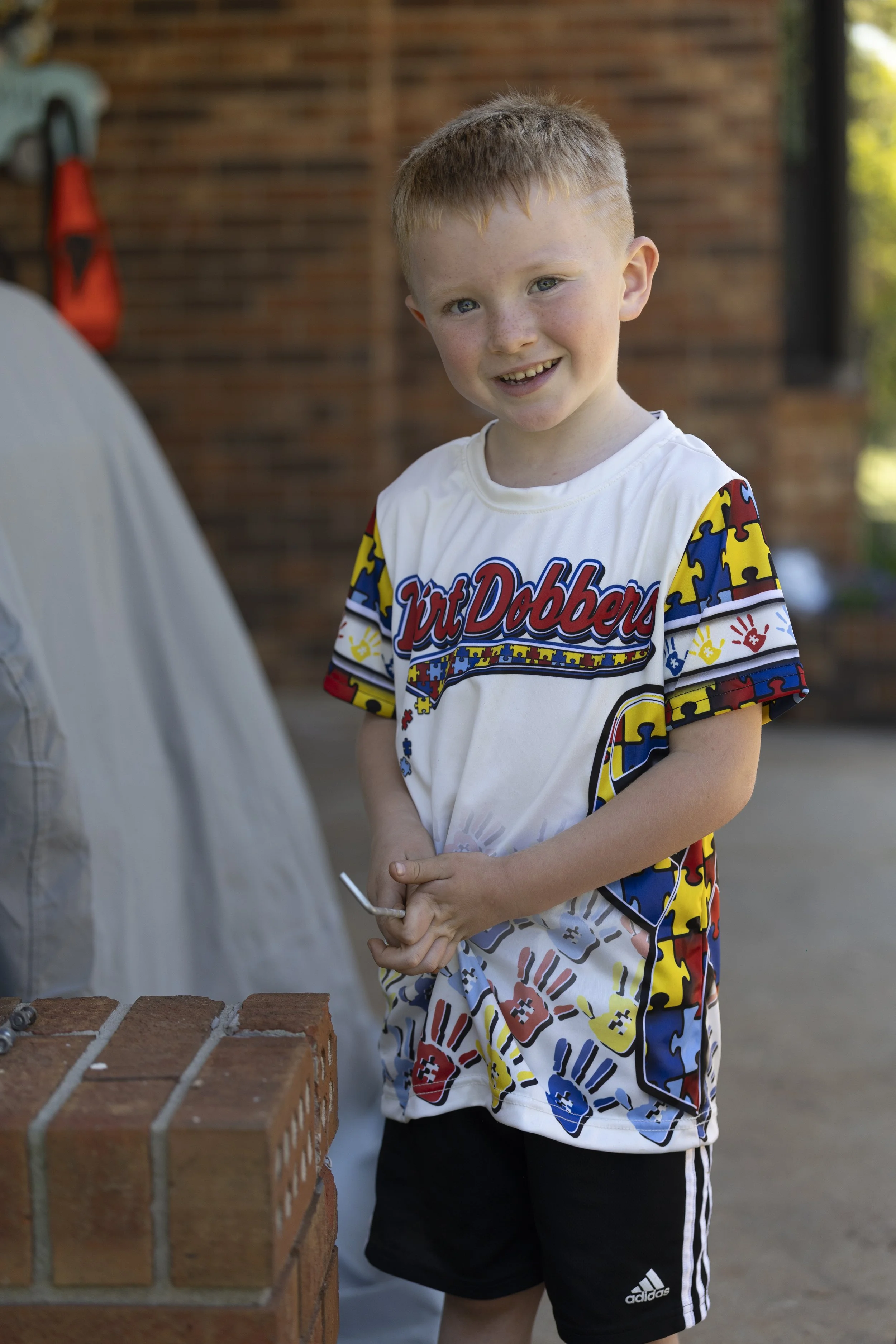 A young boy with red hair and freckles stands outdoors near a brick barbecue grill, smiling at the camera, wearing a colorful 'Toy Dobbins' t-shirt and black Adidas shorts.
