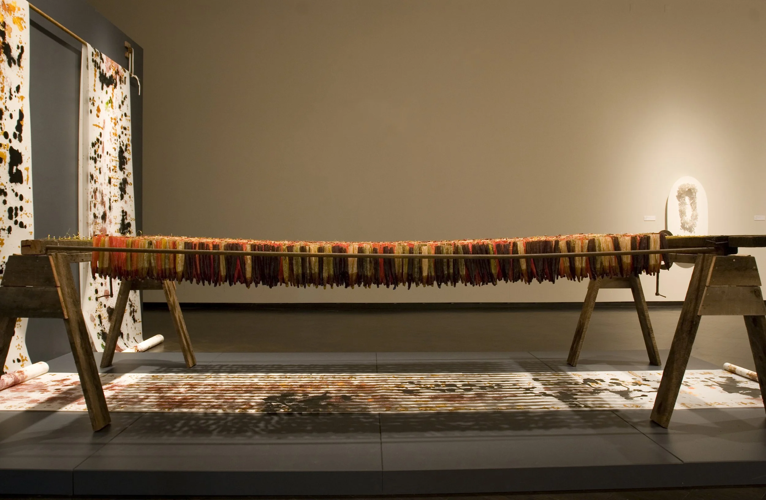 Art installation featuring a long horizontal artwork made of colorful sticks on a wooden structure, with abstract patterned panels on the left and a sign on the right in a gallery setting.