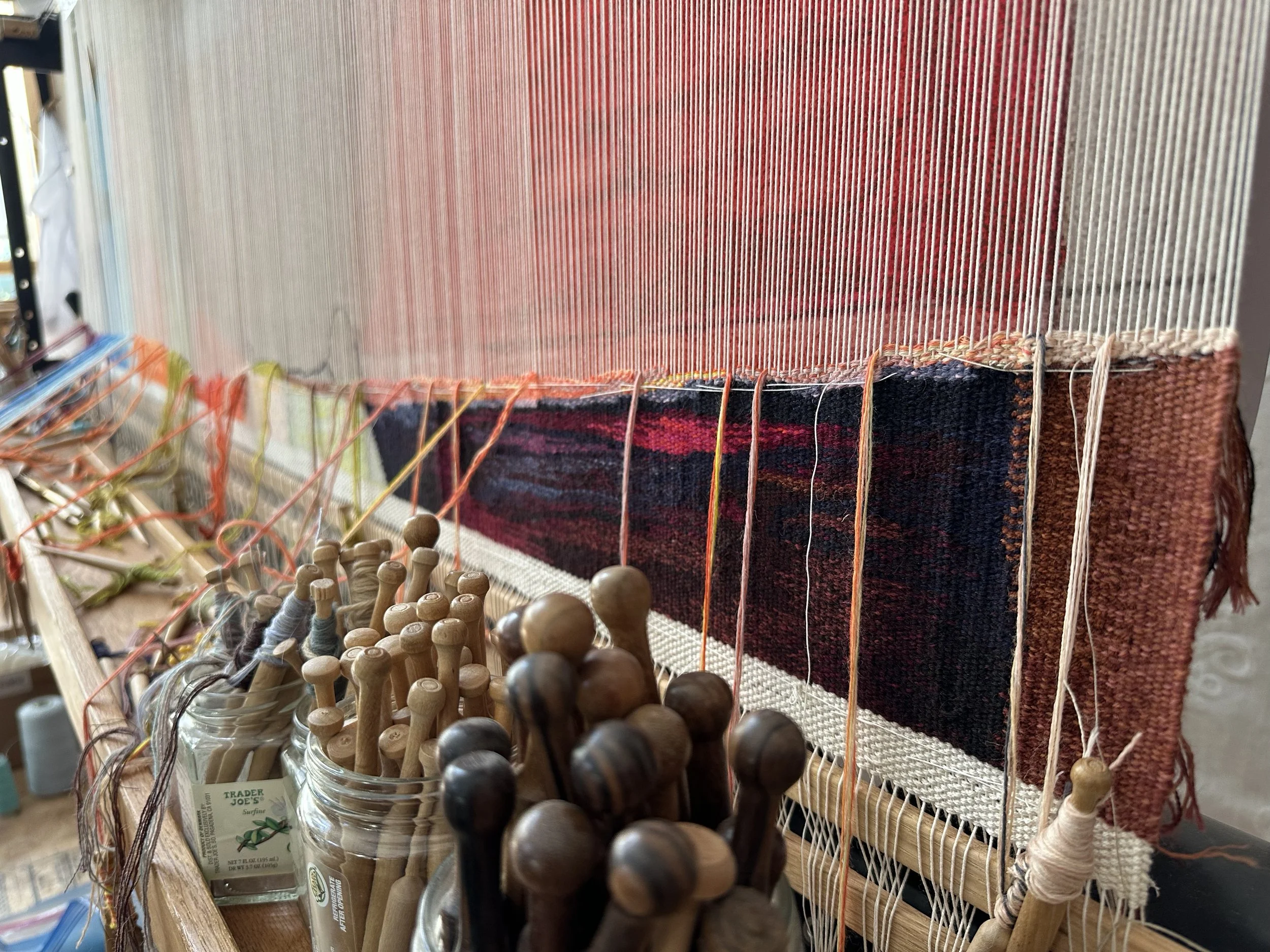 A weaving loom with colorful woven fabric and wooden bobbins in jars in the foreground.