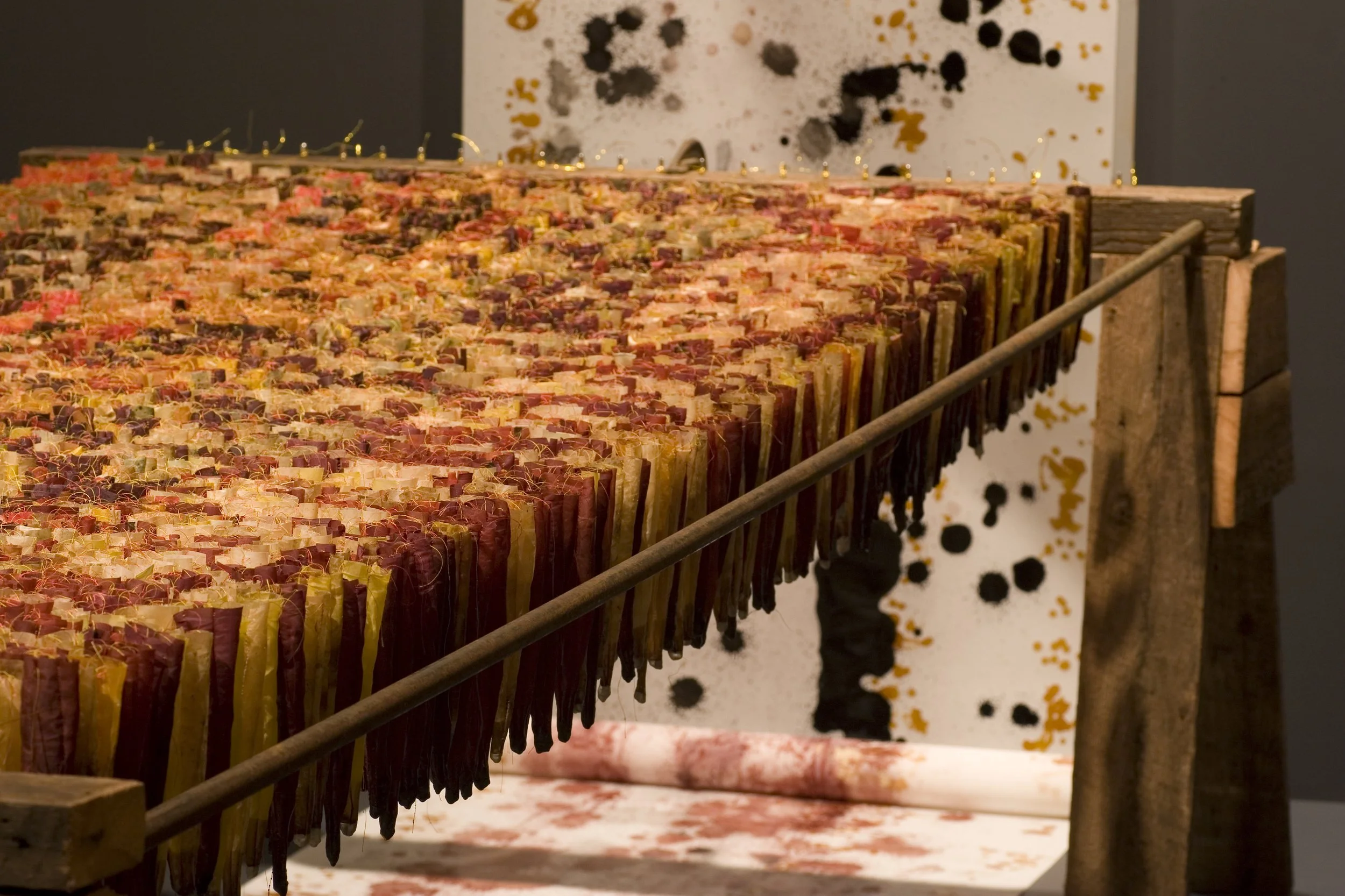Close-up of an art installation featuring a large collection of colorful, bundled objects hanging on a rack, with a background of splattered black, gold, and pink paint on a white wall.