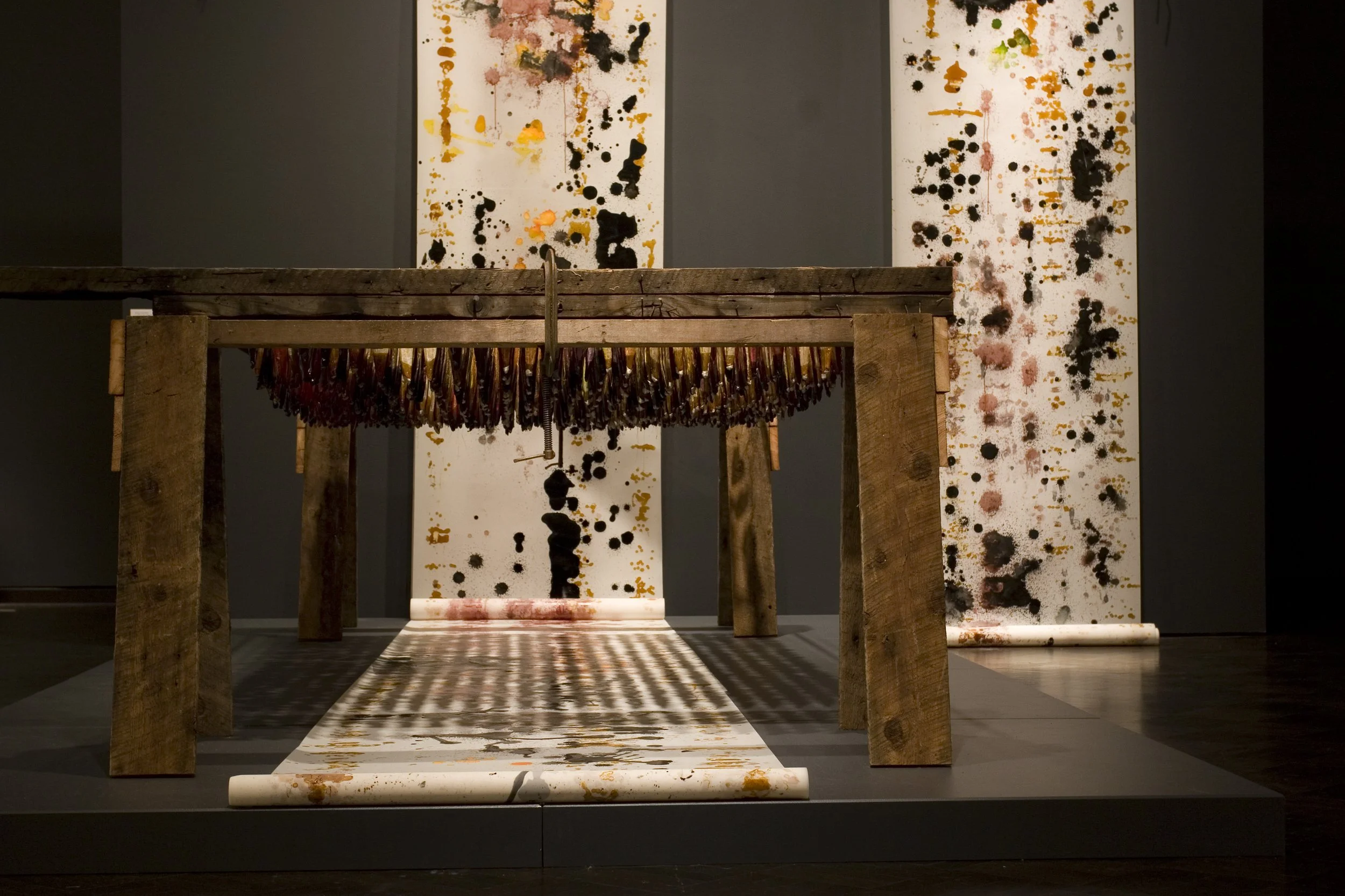 An art installation featuring two abstract paintings with black, orange, yellow, and pink splatters and drips on a white background, with a rustic wooden table and a rolled-up canvas underneath.