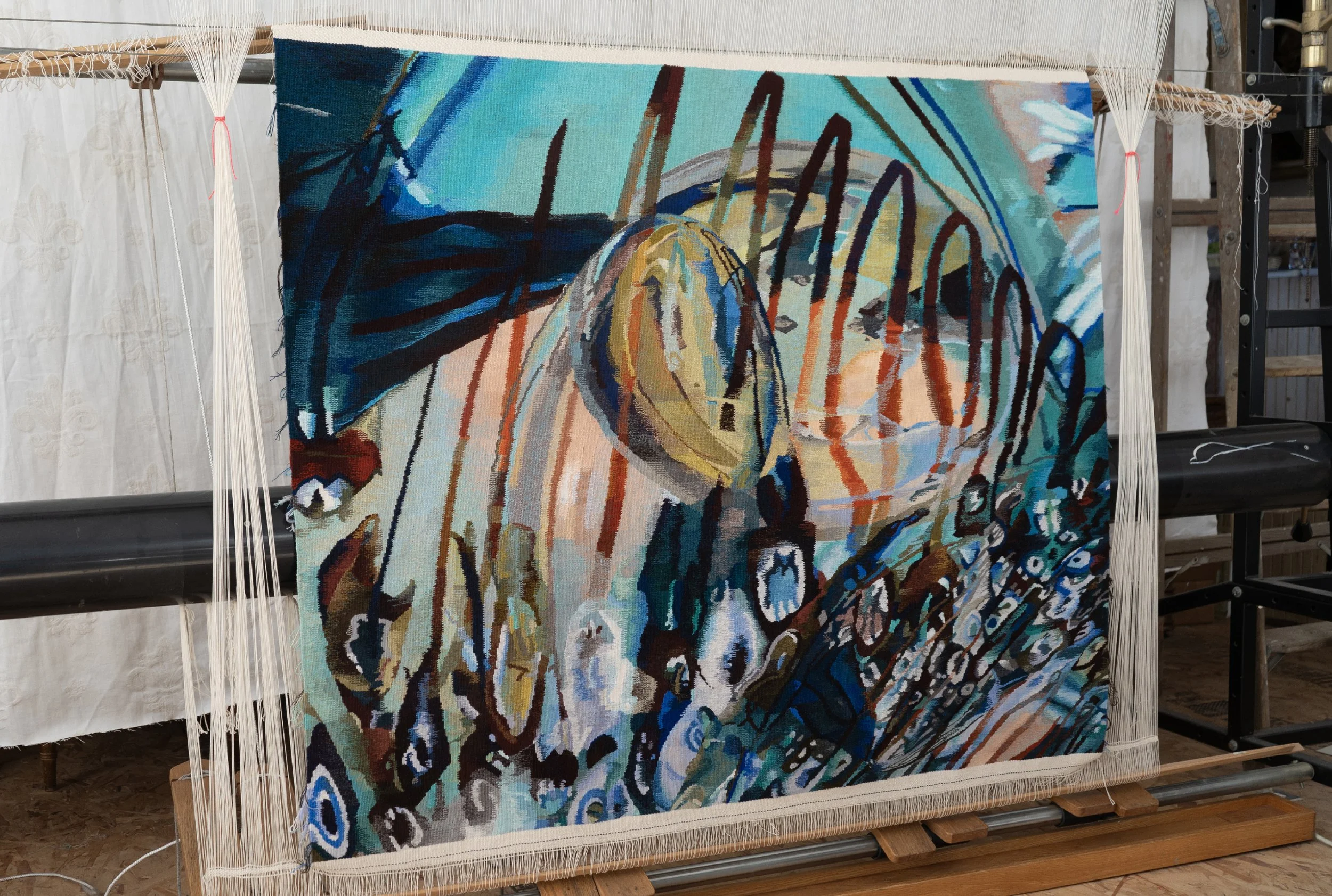 Close-up of a colorful woven tapestry with abstract waves and the word 'ALOHA' incorporated into the design, mounted on a loom.