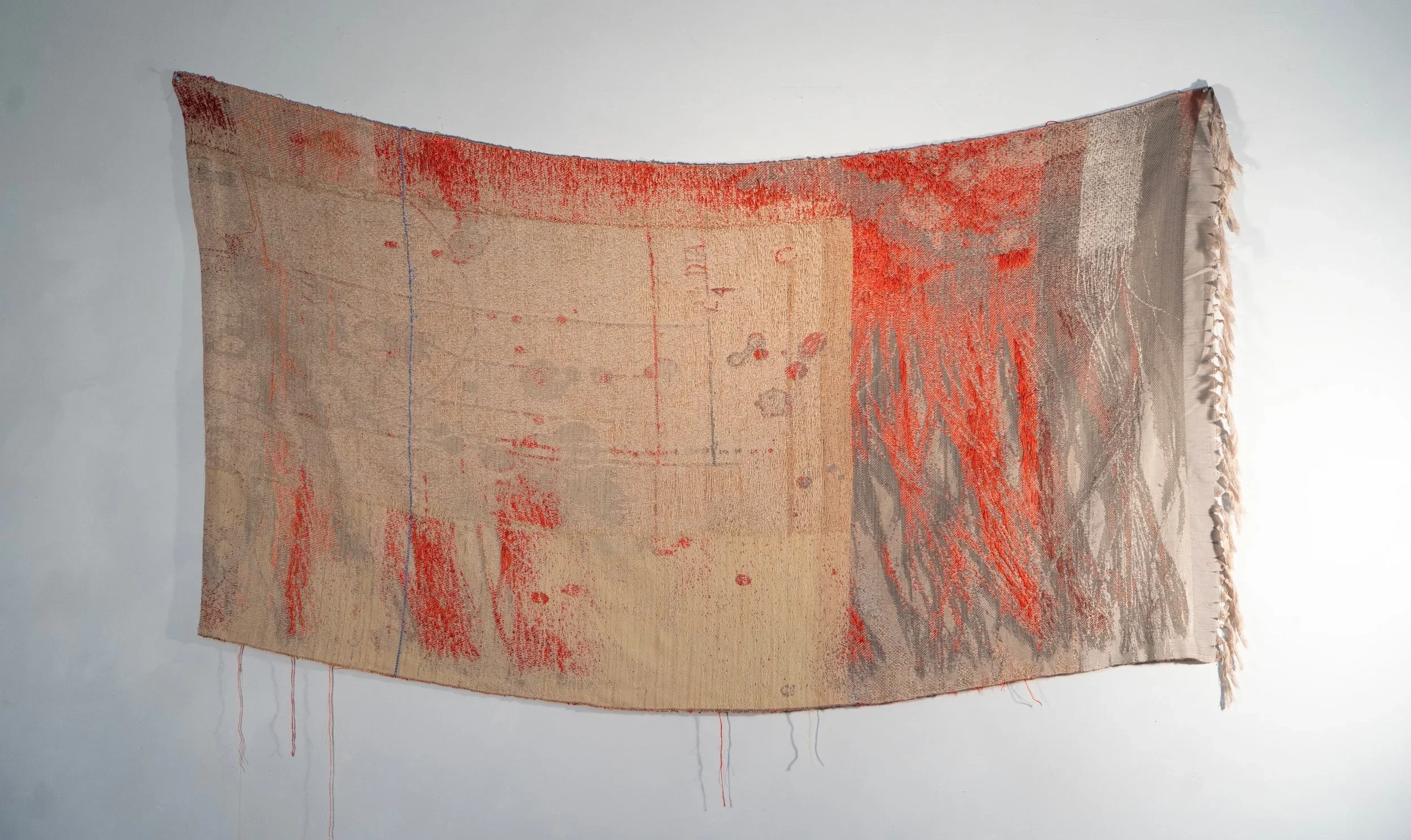 A textile artwork with abstracted geometric and organic shapes in beige, red, and gray tones, suspended on a white wall.