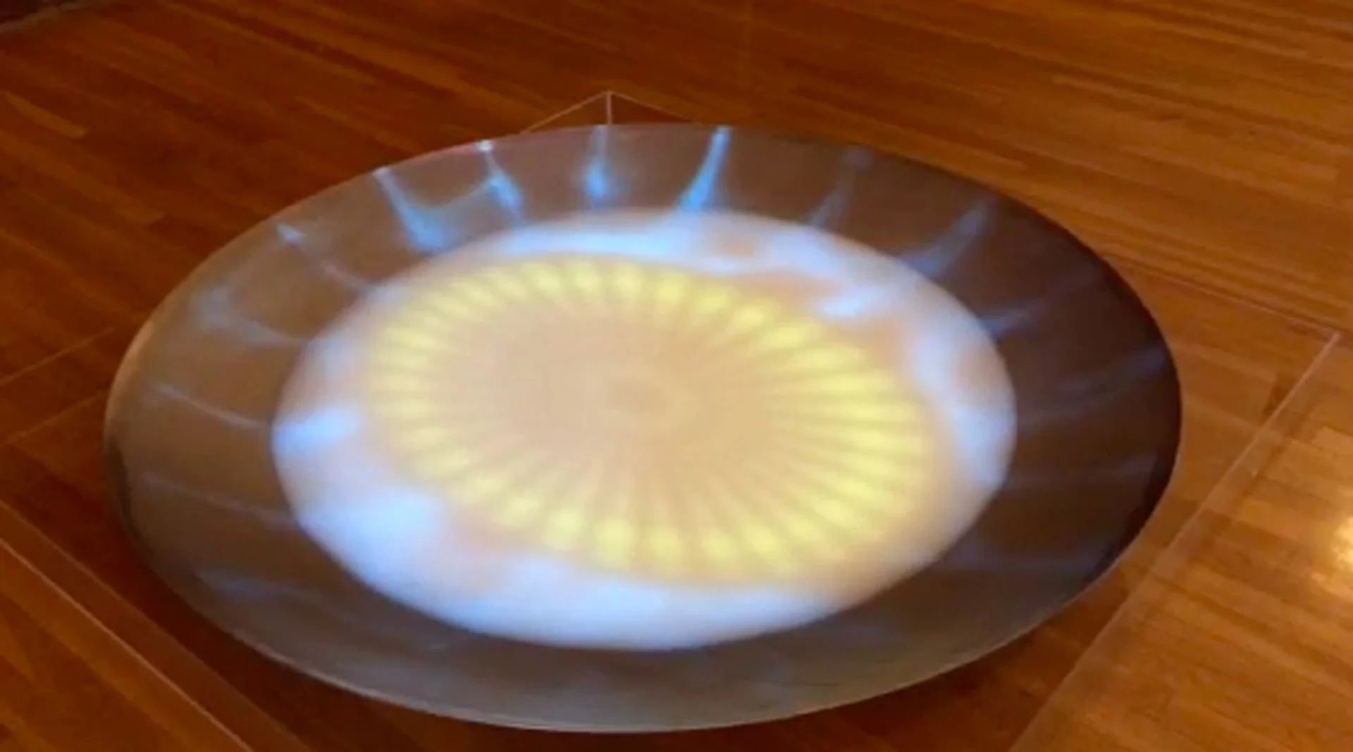 A metallic bowl on a wooden floor, reflecting a pattern of yellow and white light.