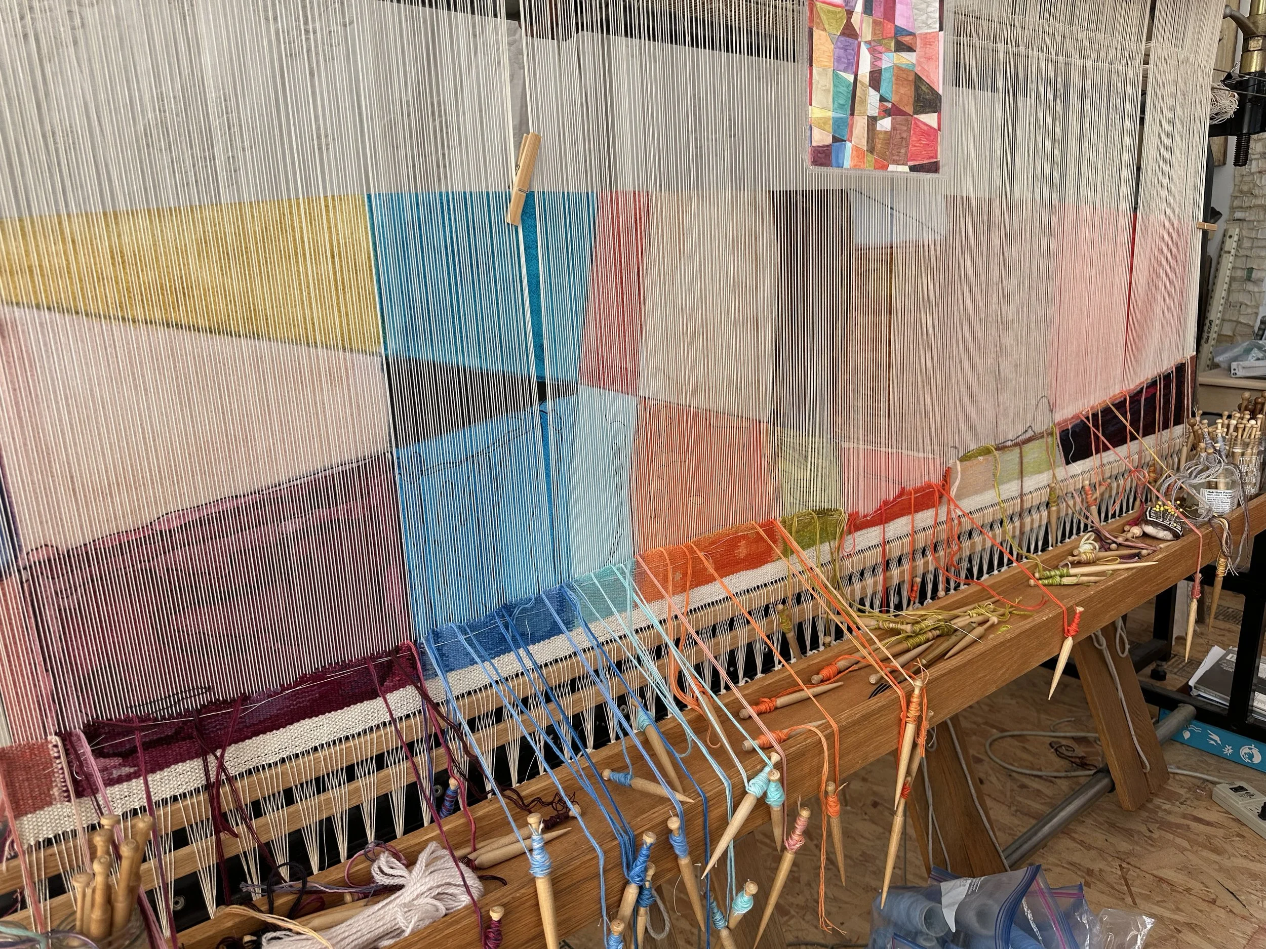 A weaving loom with colorful yarns in pink, yellow, blue, and red, and a partially woven fabric with a geometric pattern, in a craft room.