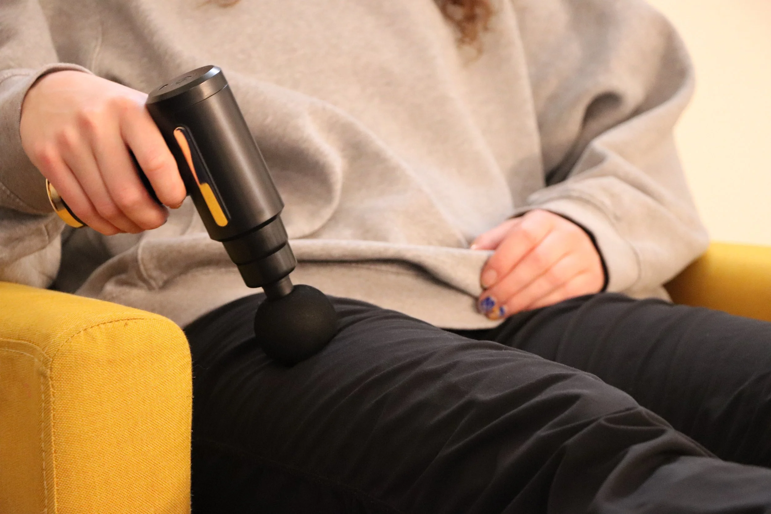 Person sitting on yellow chair using a massage gun on their thigh, wearing gray sweatshirt and black pants.
