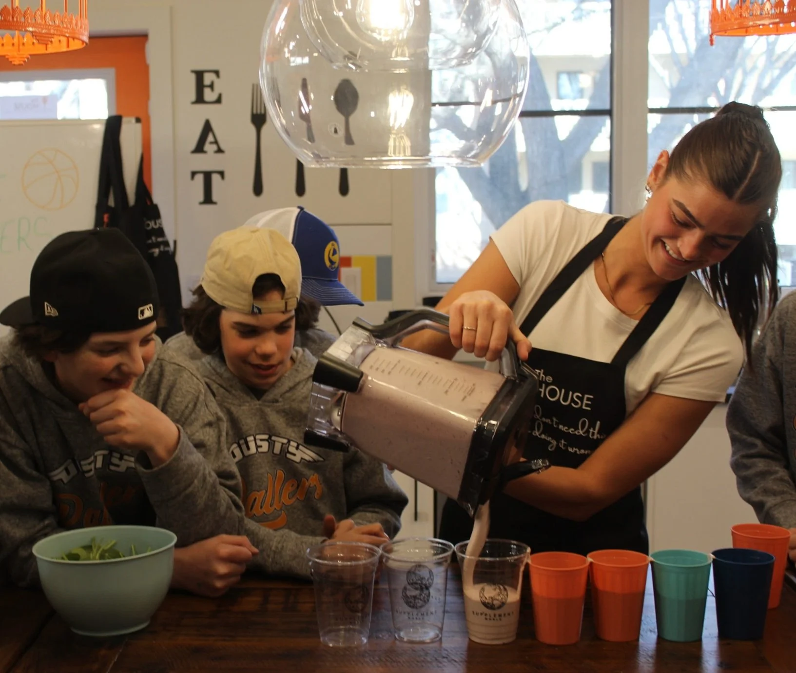 A woman in an apron pours a pink smoothie from a blender into clear cups, with three young hockey teammates watching and smiling at a wooden table in a kitchen setting.