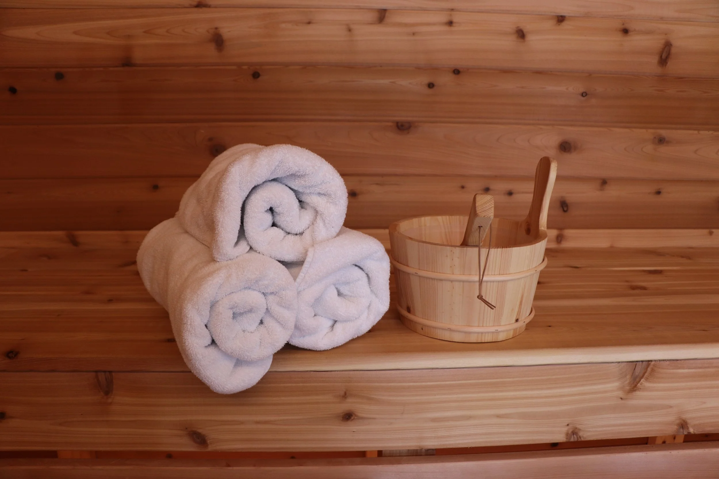 Stack of three rolled white towels next to a wooden bucket with a wooden dipper, all on a wooden surface with a wooden wall background.