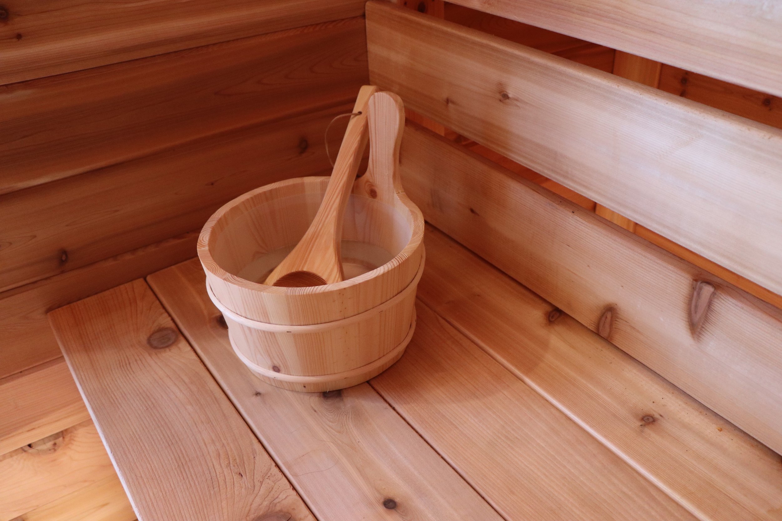 A wooden bucket with a wooden ladle hanging inside it, placed on a polished wooden surface inside a sauna with wooden walls.
