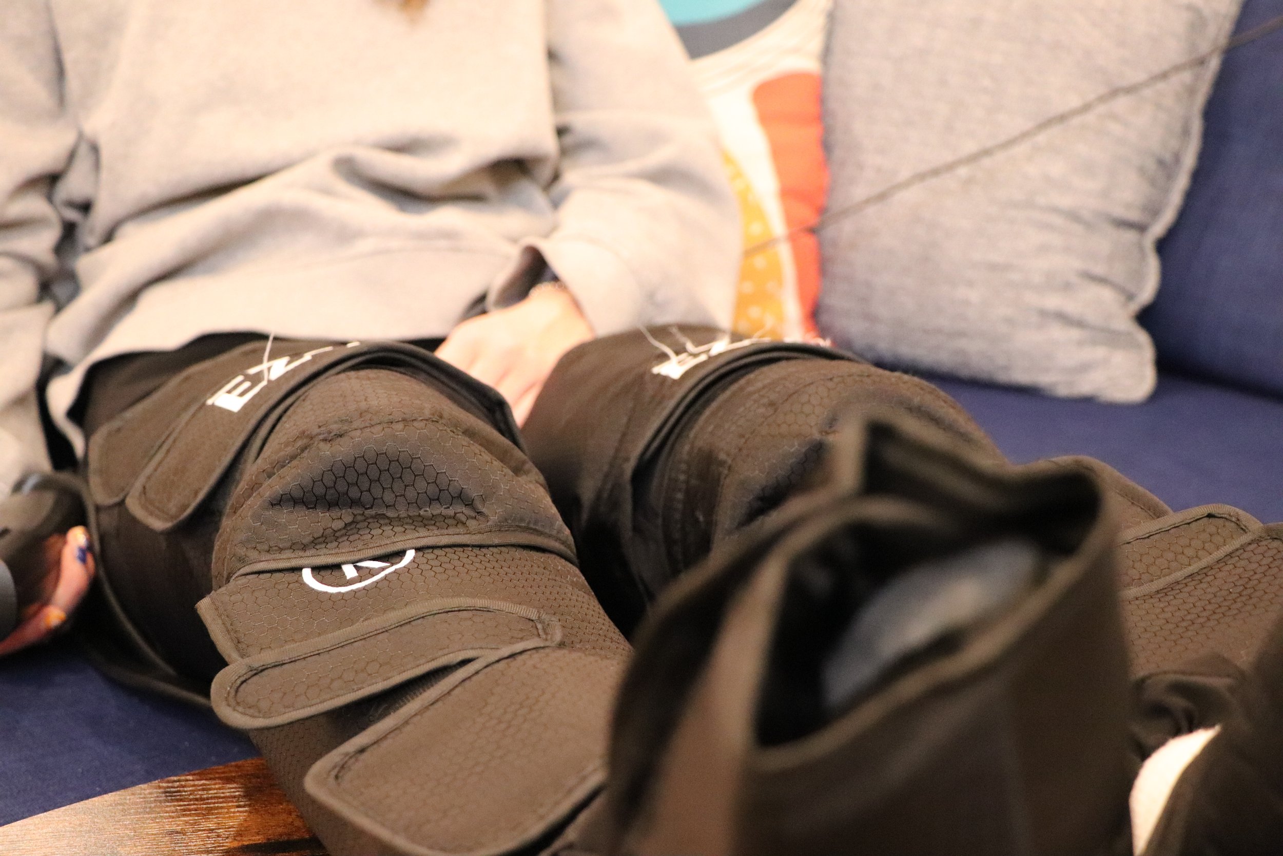 Close-up of a person's legs wearing brown hiking boots, with socks visible, and light-colored pants. The background shows part of a couch or chair with a gray blanket or clothing draped over it.