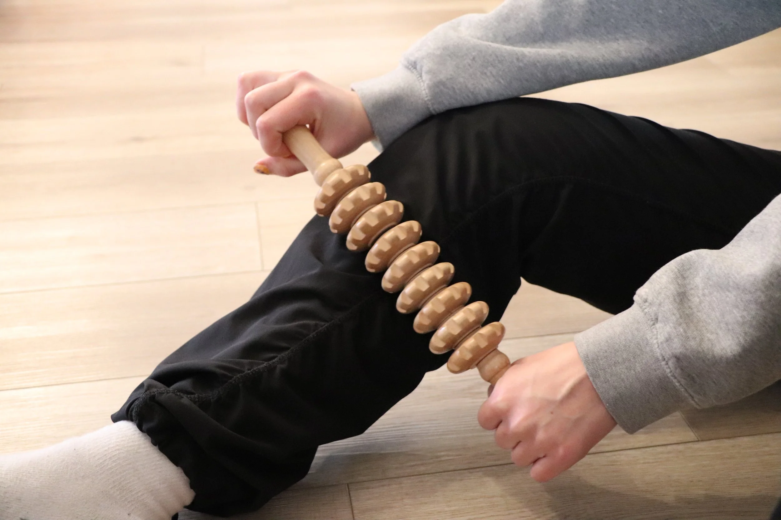 Person sitting on the floor wearing black pants and a gray sweatshirt, holding a wooden massage roller on their leg, with a hand assisting gently.