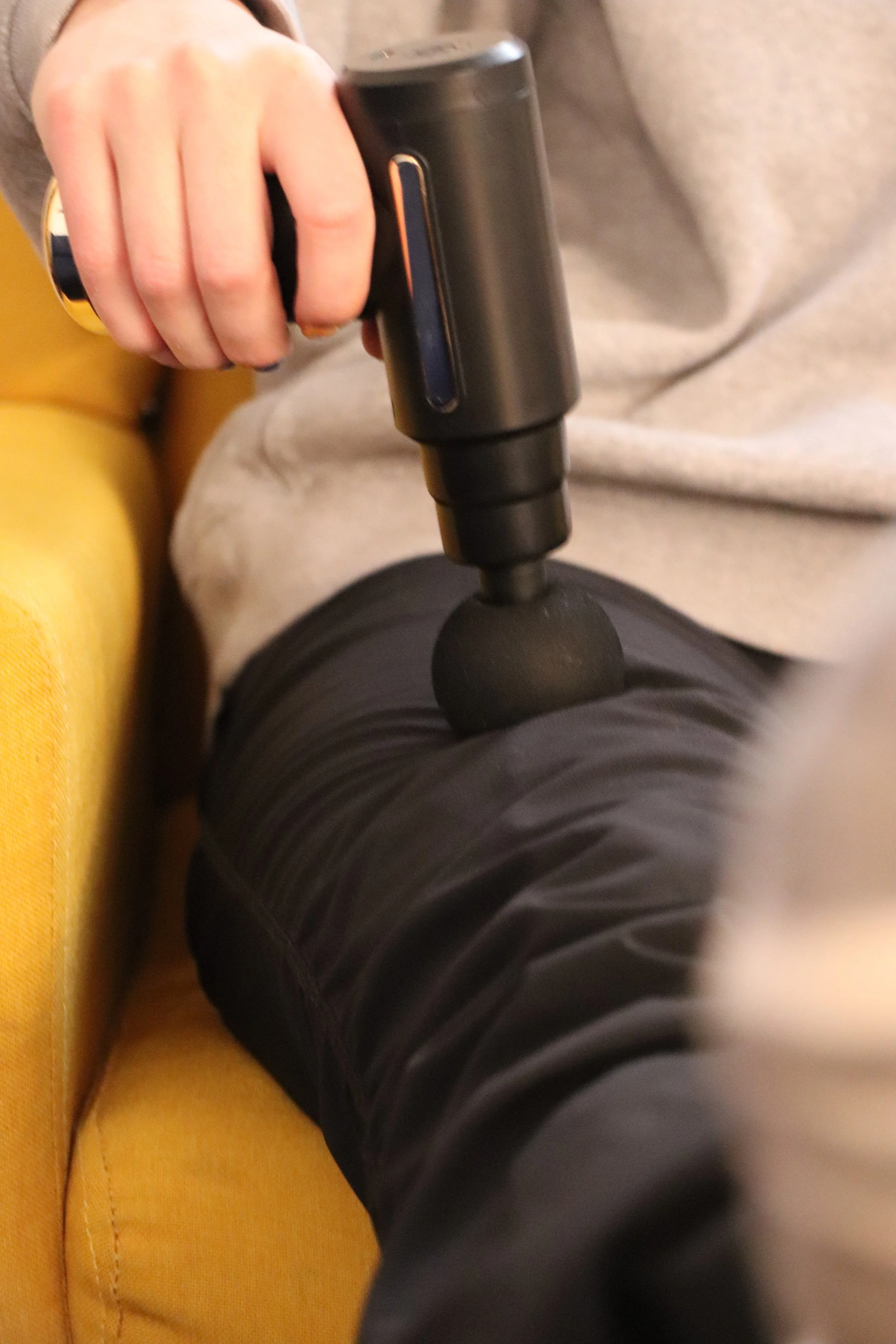 Person holding a massage gun on their thigh, wearing black leggings and a grey top, sitting on a yellow couch.