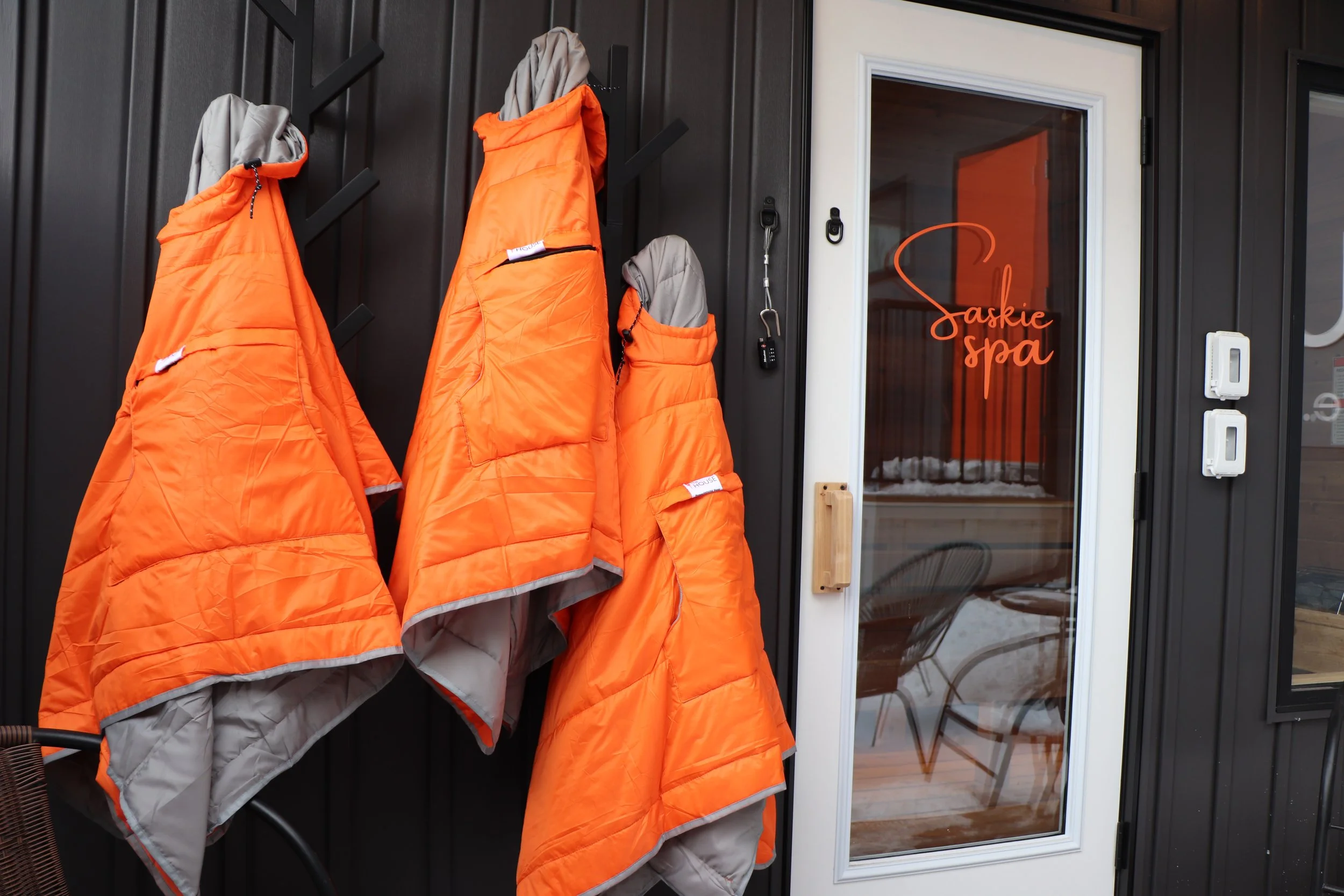 Four orange and gray winter jackets hanging on black hooks outside of a spa called Saskie Spa.