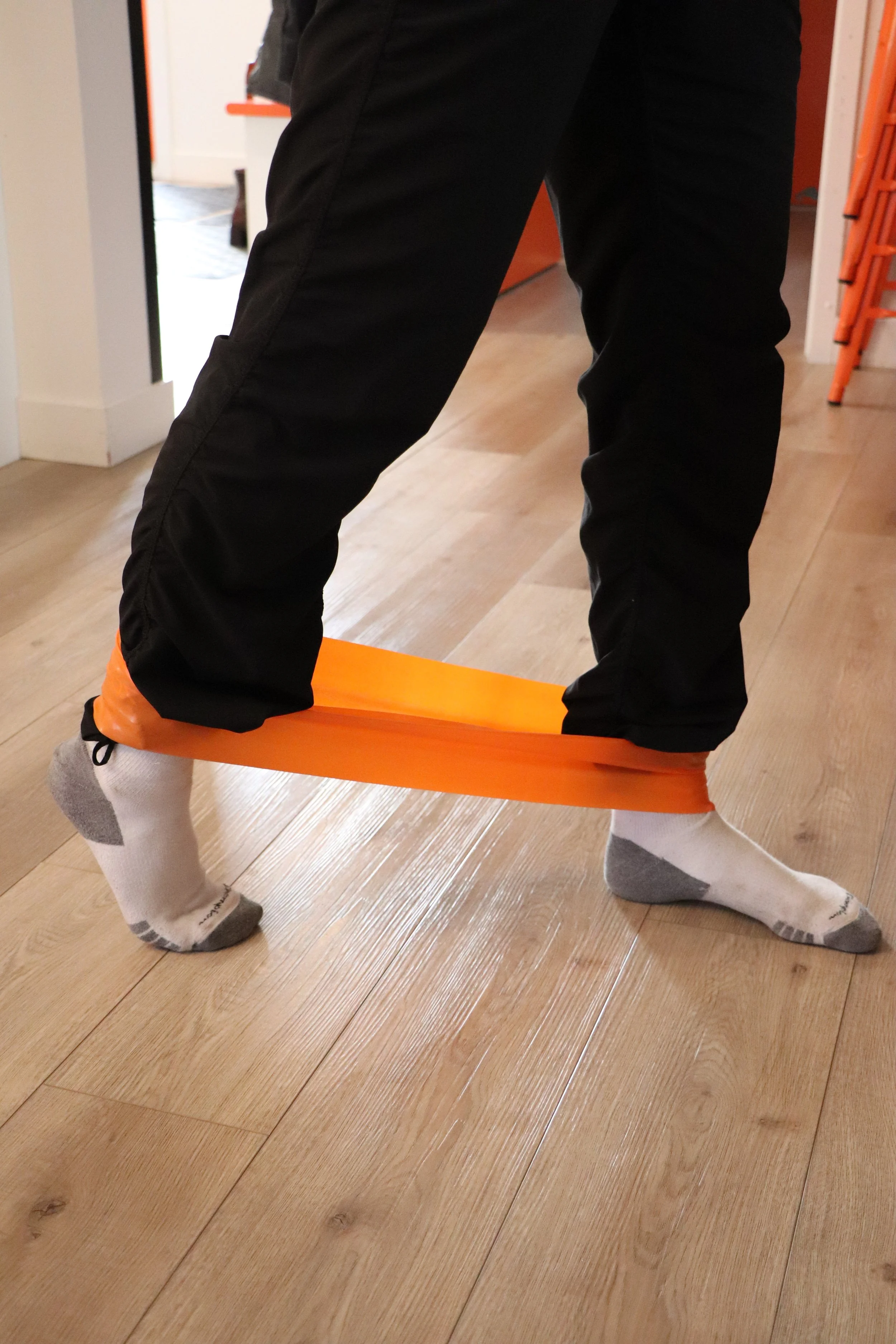 Person standing with one foot on an orange balance board, wearing black pants, orange socks, and gray and white socks, on a hardwood floor in a room with orange and white walls.