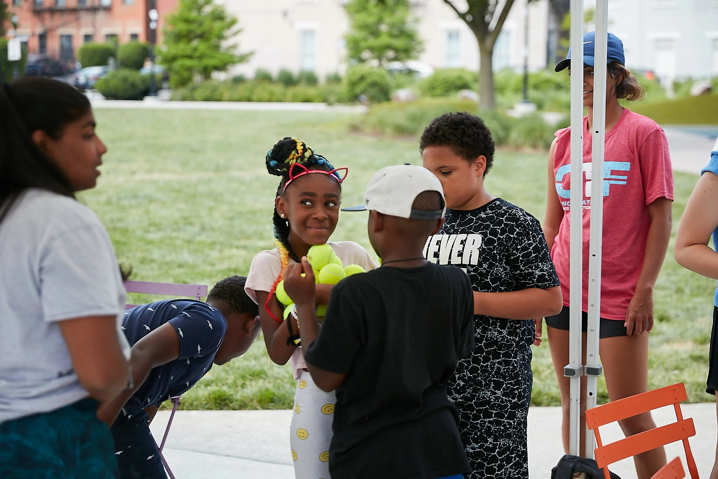 Children gathered around a girl holding several tennis balls at an outdoor event in a park.