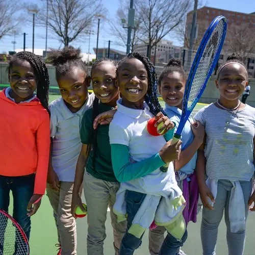 Group of six young girls outdoors on a tennis court, smiling, with one holding a tennis racket and a ball, dressed in casual sportswear.