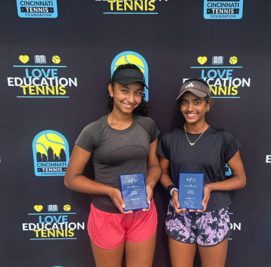 Two young girls holding awards in front of a black backdrop with tennis and education logos, smiling at a tennis event.