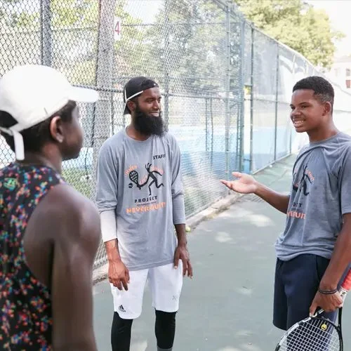 Three people talking and smiling on a tennis court, with a chain-link fence in the background.