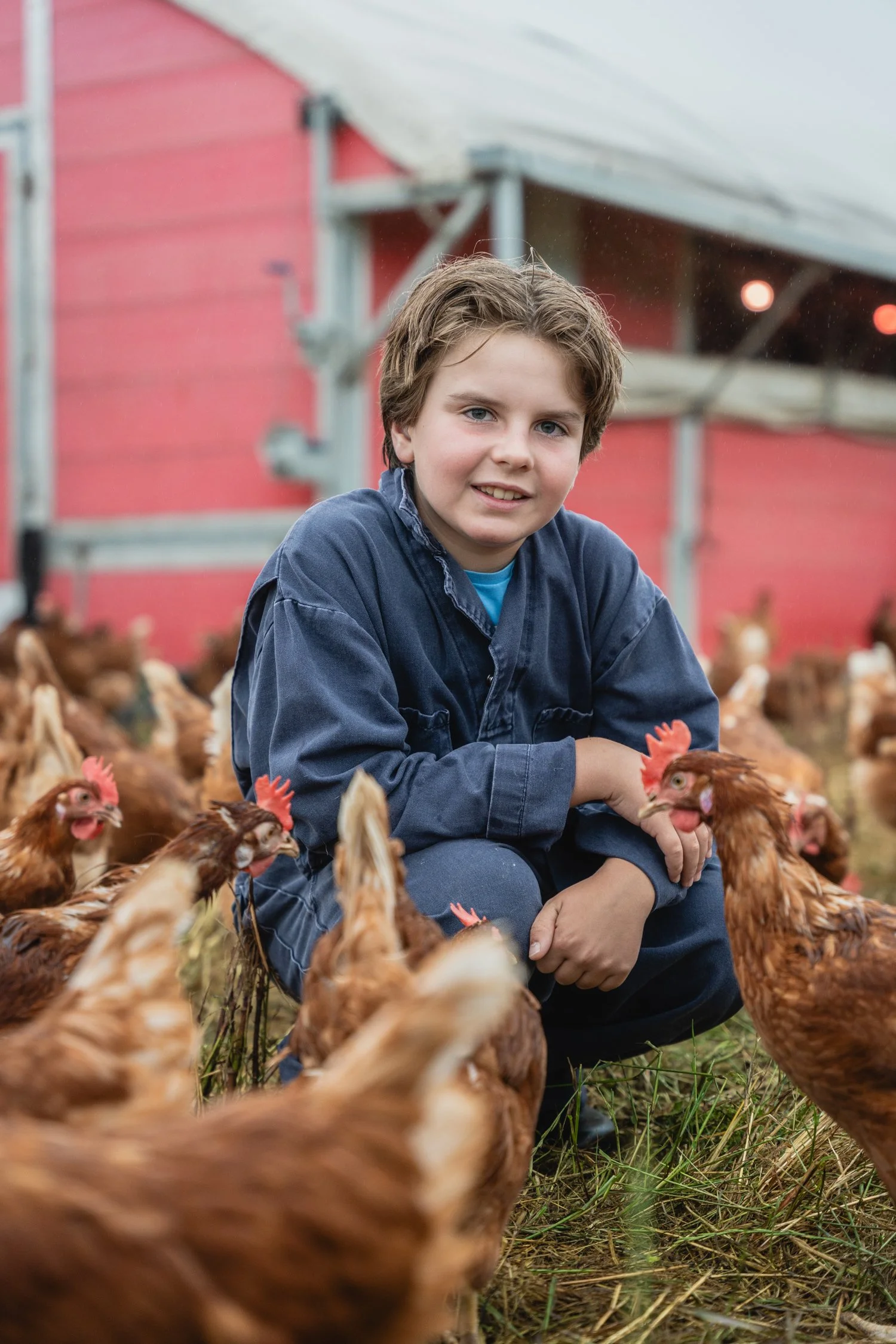 Charlie Wilcox, fifth-generation Wilcox family member, crouching with pasture-raised chickens in front of a farm barn.