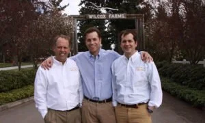 Three men standing outdoors in front of a farm sign, smiling with arms around each other, wearing business casual attire.