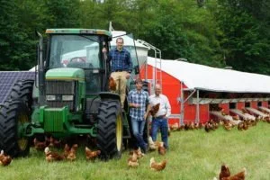 Group of people standing next to a large green tractor and a chicken coop, with chickens on the ground, in a farm setting.