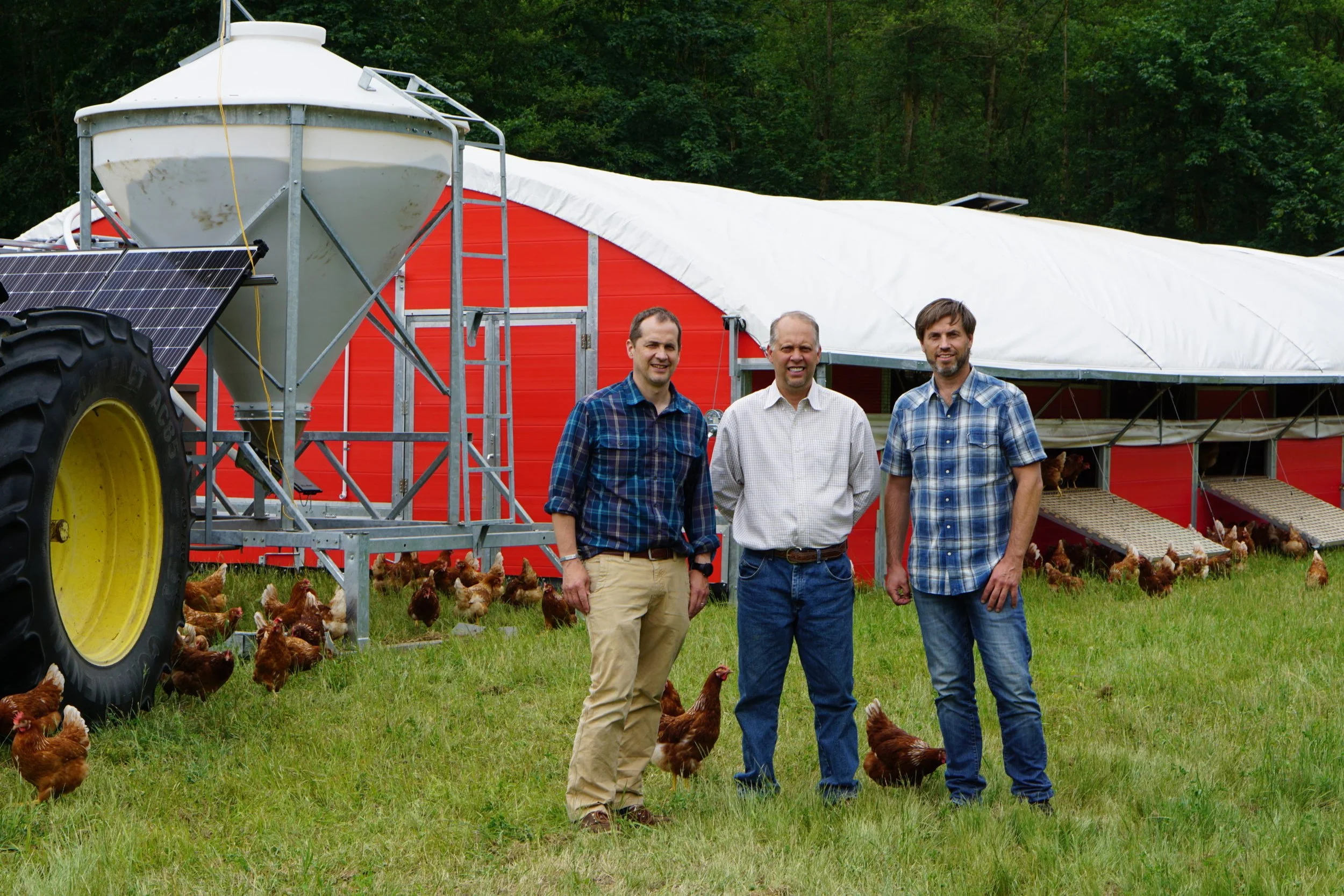 Three men standing on a farm with chickens, a red barn, and a tractor with solar panels in the background.
