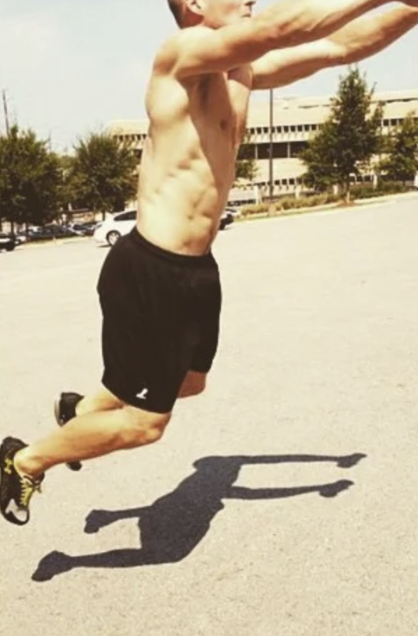 A shirtless man jumping on an outdoor parking lot with his arms extended forward, casting a shadow on the ground.