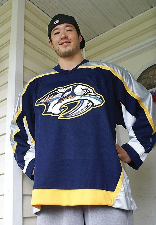 A young man wearing a Nashville Predators hockey jersey and a black baseball cap, standing outside a house, smiling at the camera.