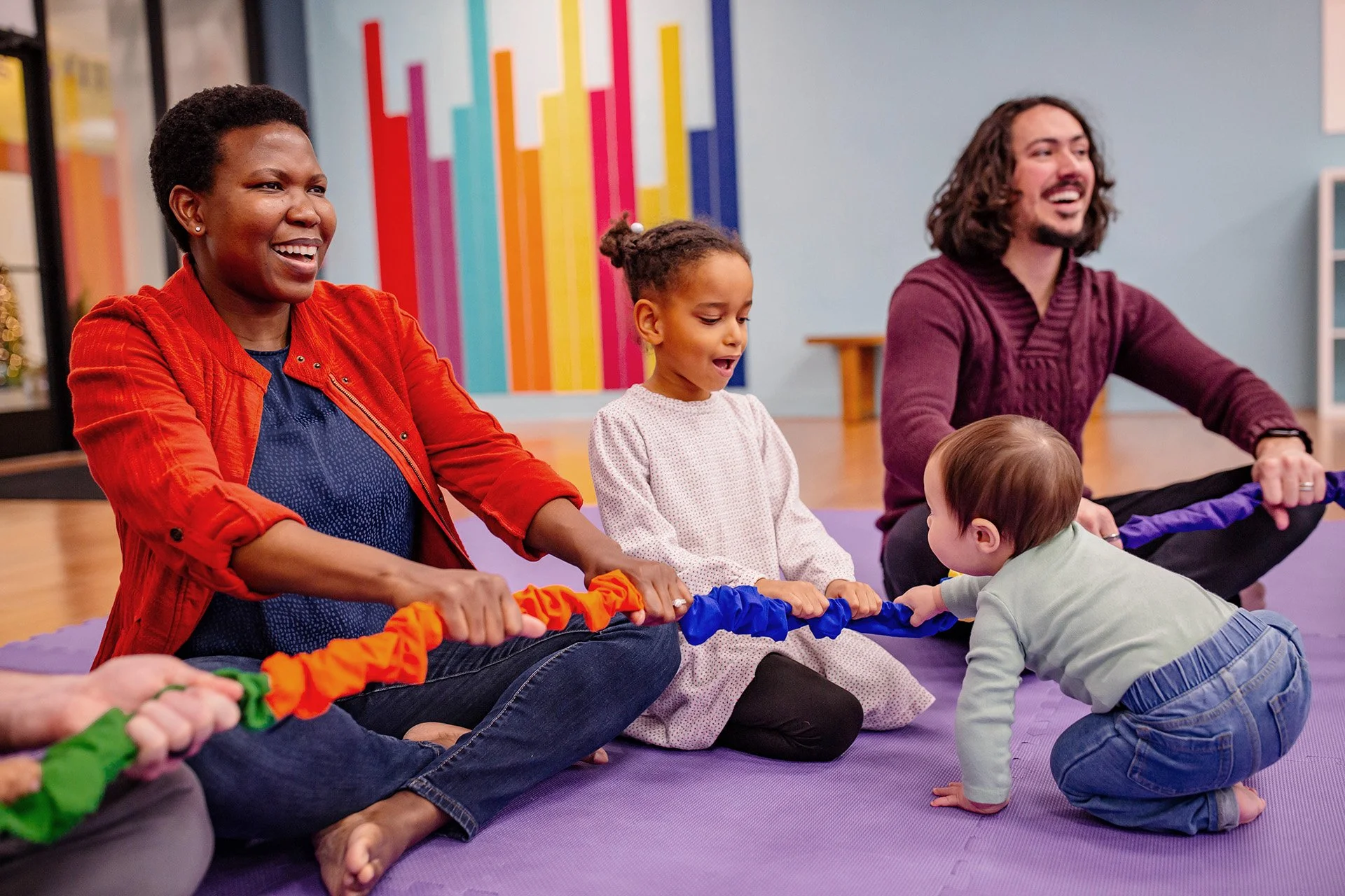 A group of adults and children participating in a group activity, sitting on a purple yoga mat, holding a colorful rope, with a colorful art piece on the wall behind them.
