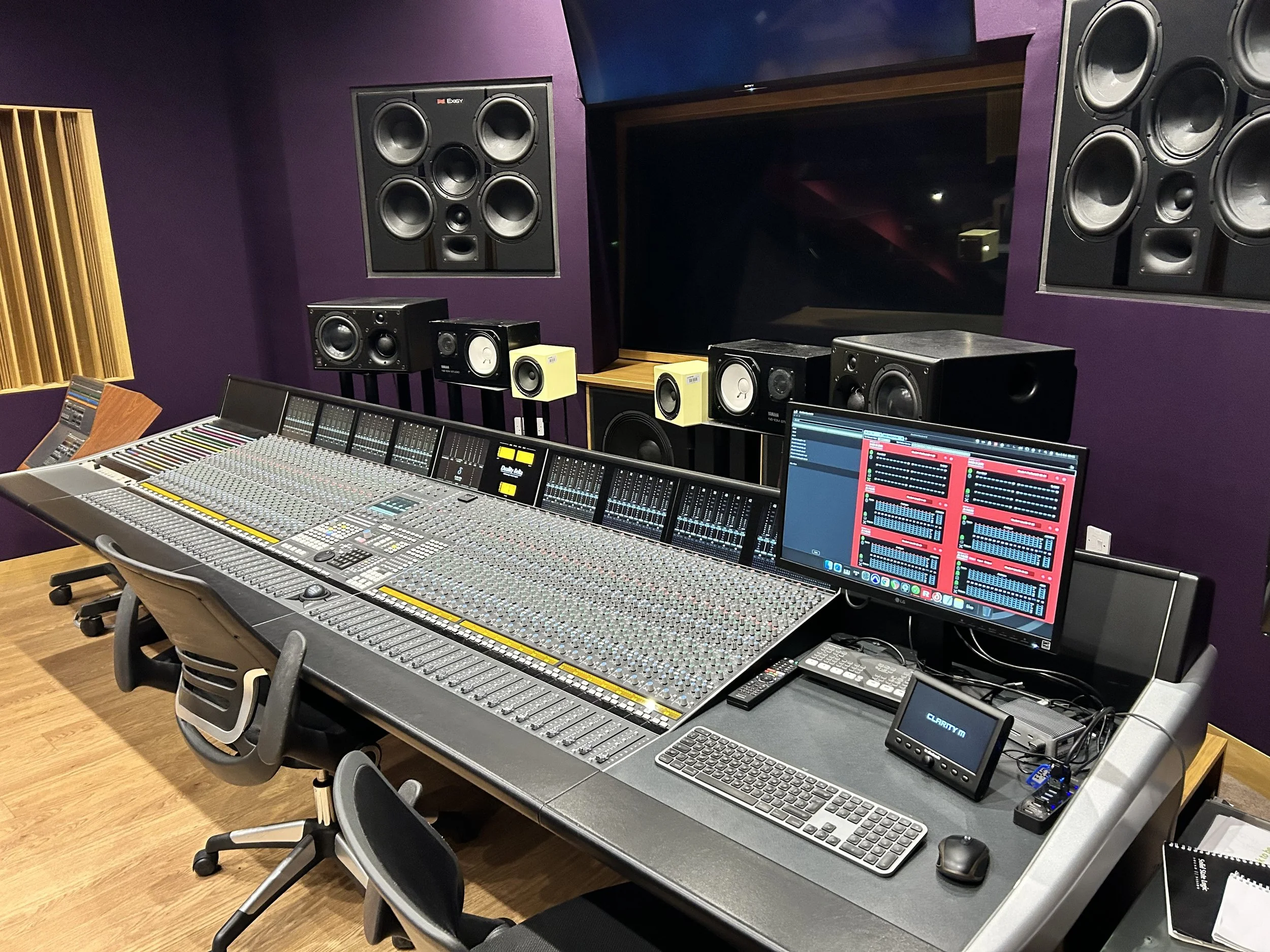 A professional recording studio control room featuring a large mixing console, studio monitors, and a computer screen with music production software, with purple walls and wooden flooring.